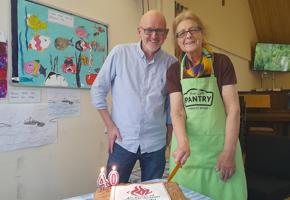Woman in a green apron holding a knife to a white cake with Church action poverty logo
