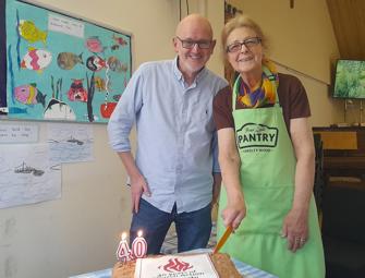 Woman in a green apron holding a knife to a white cake with Church action poverty logo