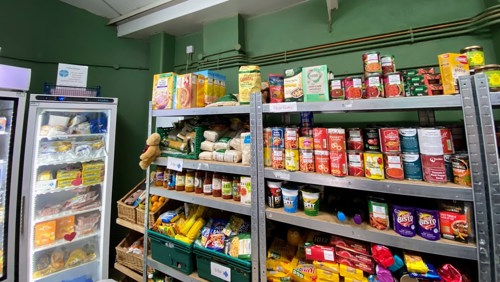Shelves stacked with canned and packet food alongside a glass fronted fridge and freezer