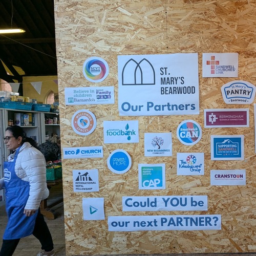 Notice board with Pantry partners and a volunteer, with shelves full of food behind her