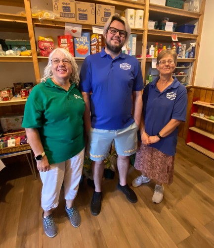 Three volunteers in Pantry Polos shirts stand and smile with shelves full of food behind