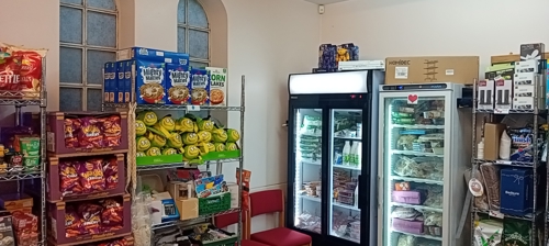 Glass fronted fridge and freezer stocked with food, alongside  shelves stocked with food and other essentials