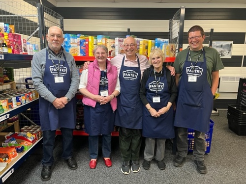A group of vounteers in Pantry aprons stand in front of shelves stocked with food.
