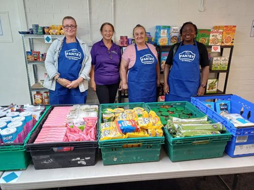 A group of vounteers in Pantry aprons stand in front of shelves stocked with food.