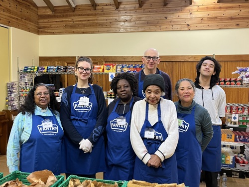 Group of smiling people in Pantry aprons with shelves of packet and canned food behind