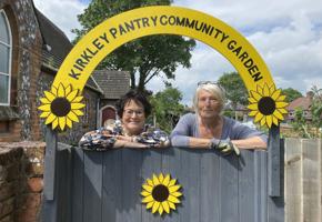 two woman in front of Kirkley Pantry Community Garden sign with sunflowers on front of grey fence