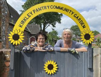 two woman in front of Kirkley Pantry Community Garden sign with sunflowers on front of grey fence