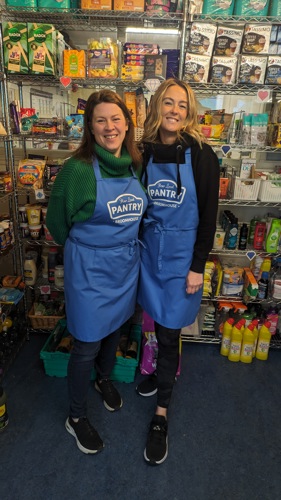 Two ladies in Pantry aprons in front of a shelf of food