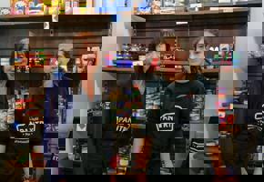 two woman in black aprons in front of food shelves 