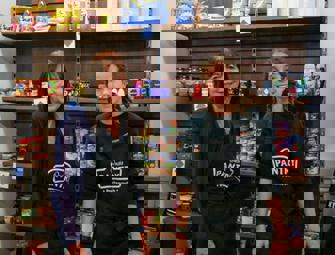 two woman in black aprons in front of food shelves 